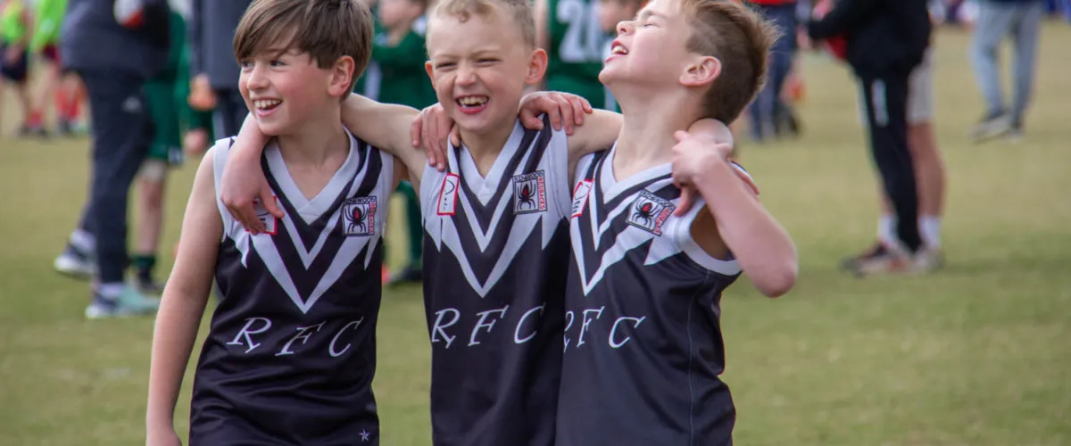 Three young Ringwood Redbacks players in club jumpers smiling with arms around each other on the field